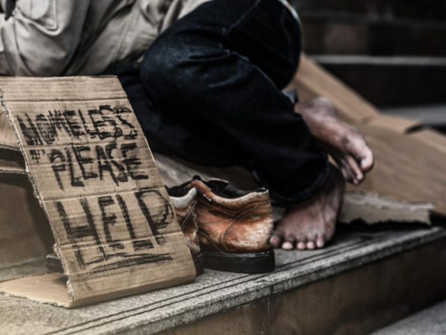 A man lies on some steps next to a sign saying "Homeless - please help"