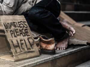 a homeless person of colour lies on some steps by a sign reading "homeless - please help"