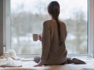 a woman sits on a bed holding a mug and looking out of a window with her back to us
