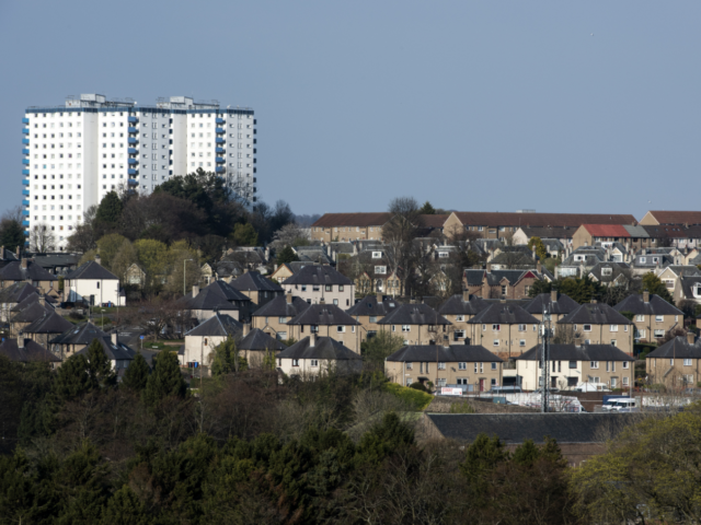 A view across a town with tower blocks in the background and houses in the foreground. There are trees among the buildings, and a blue sky