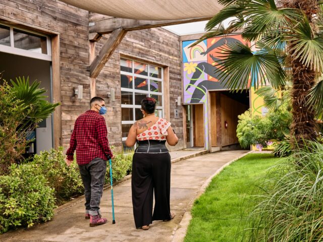 Two masked disabled people of color chat and walk through an outdoor courtyard, photographed from the back. One walks with a cane.