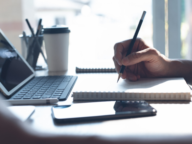 Photo of hand writing on a notebook in front of a laptop